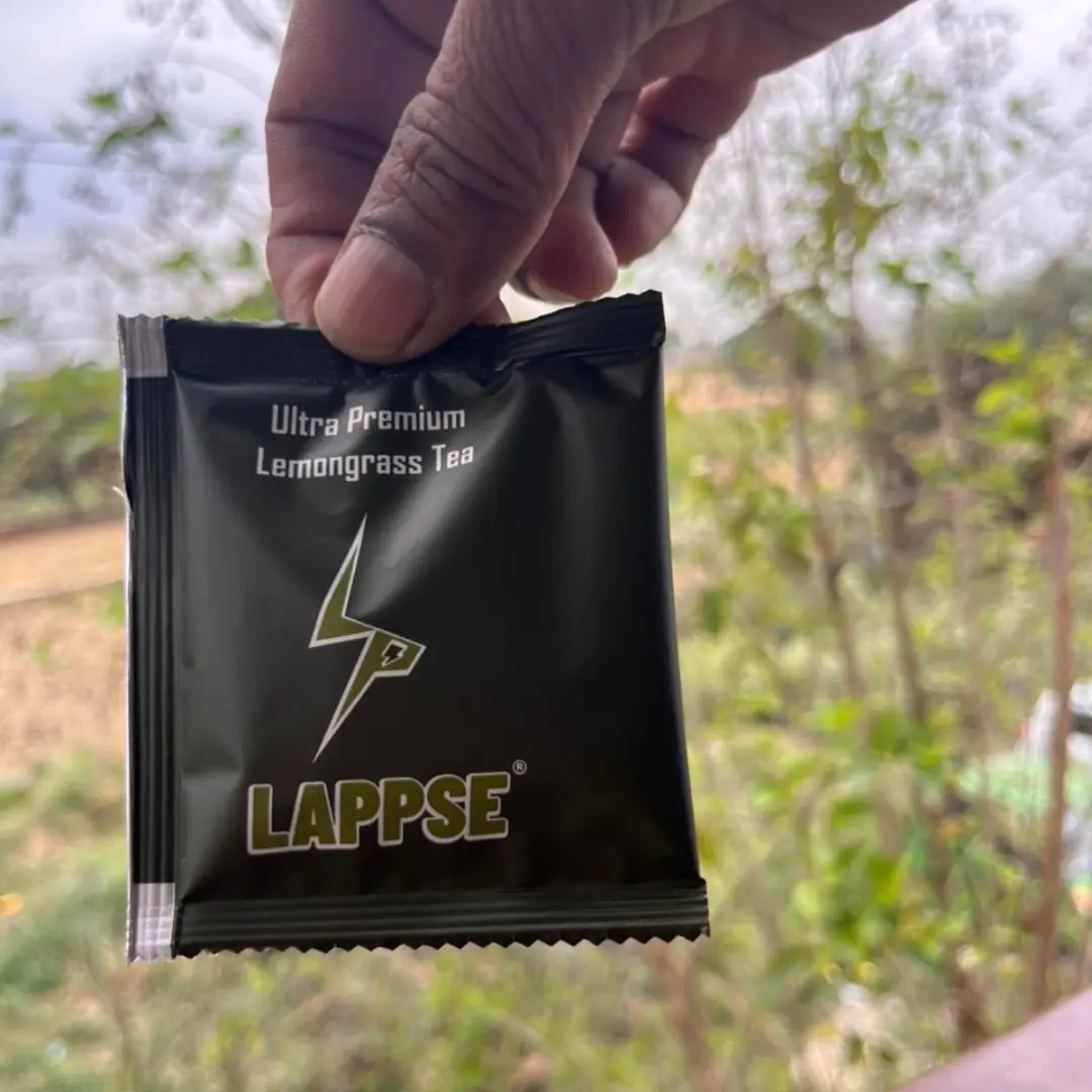 A close-up shot of a hand holding a single-serving black sachet of Lappse Ultra Premium Lemongrass Tea, featuring a sharp green and white lightning bolt logo, against a blurred outdoor background of green trees and plants.